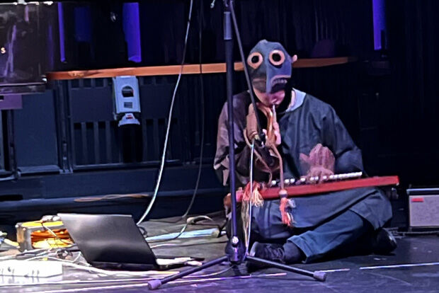 Person sat cross-legged on the floor, wearing a plague doctor's mask, playing the bugle and an electronic keyboard