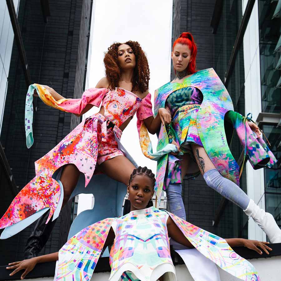 Three models wearing vibrant, futuristic, geometrically patterned fashion designs against a backdrop of modern glass buildings.