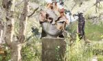 A detailed stone sculpture of a face, with natural elements like leaves and branches integrated into its design, standing on a pedestal in a sun-dappled, overgrown forest.