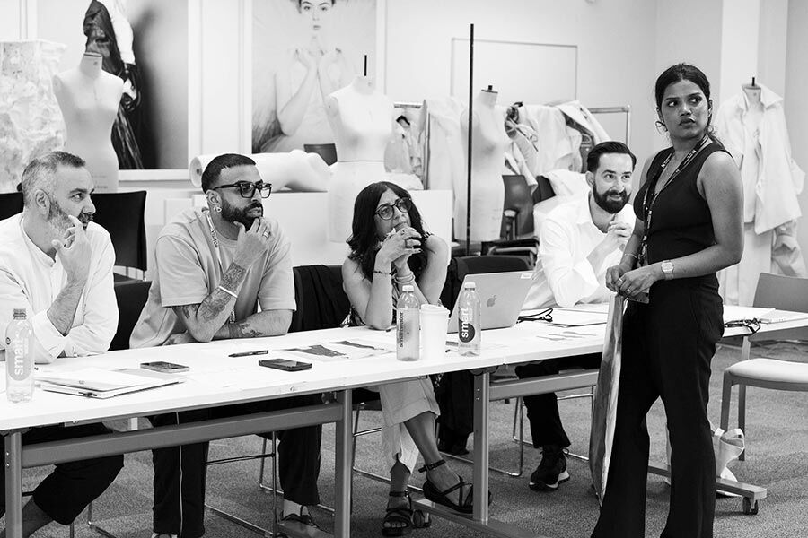 A black and white photo of five individuals, four seated and one standing, around a long table.