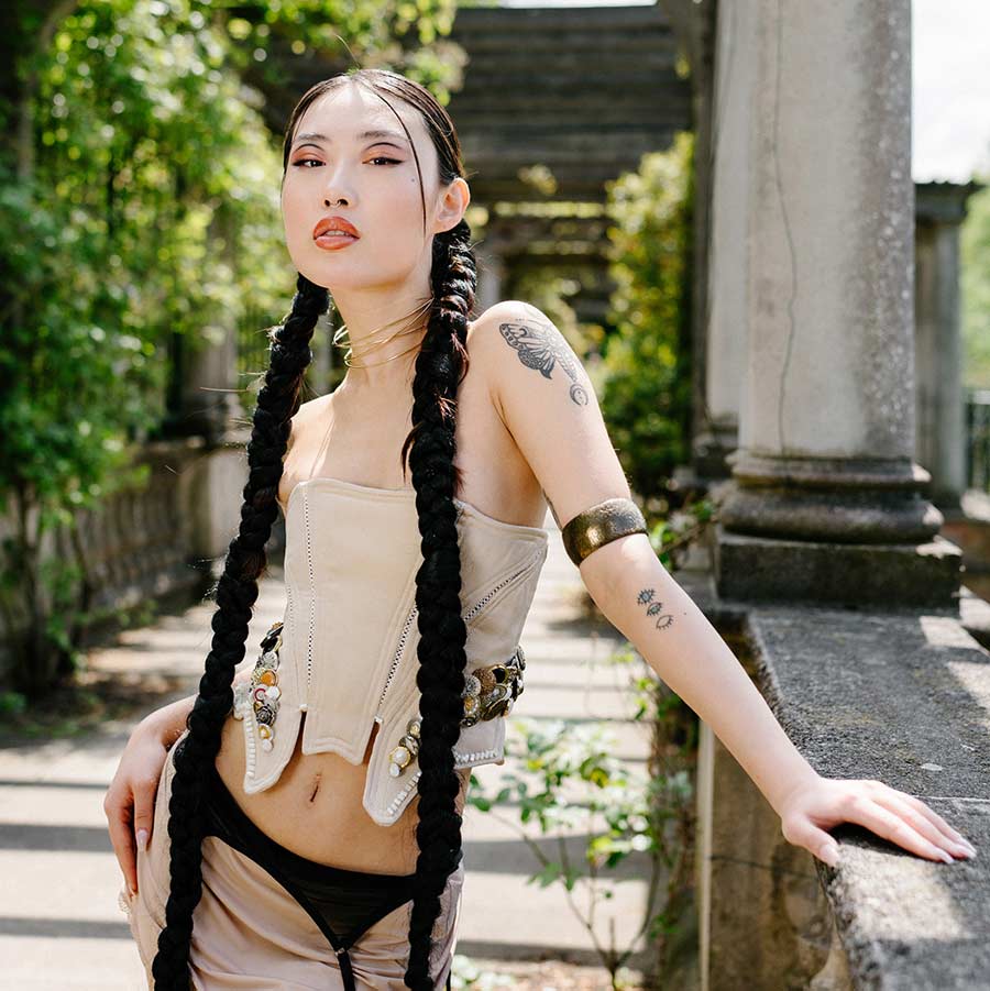 A model in a beige corset and long black braids poses in a sunny outdoor setting with stone columns and greenery.