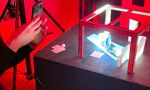 A person in a black long-sleeved shirt is holding a smartphone to photograph a small, illuminated diorama inside a red-framed box, all bathed in red light.