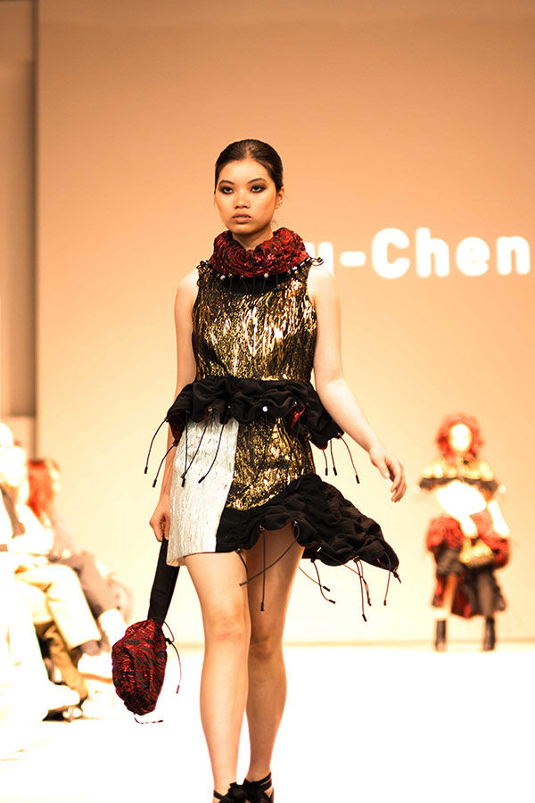 A model walks the runway in a short, metallic gold and black textured dress with a high, red ruffled neckline and an abstract black ruffle detail around the waist.