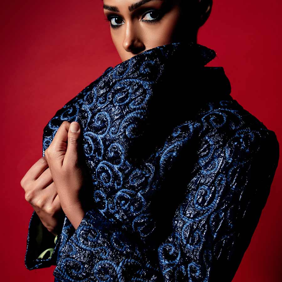 Close-up shot of a model in a dark blue, highly textured jacket with a scroll pattern, set against a vibrant red background.