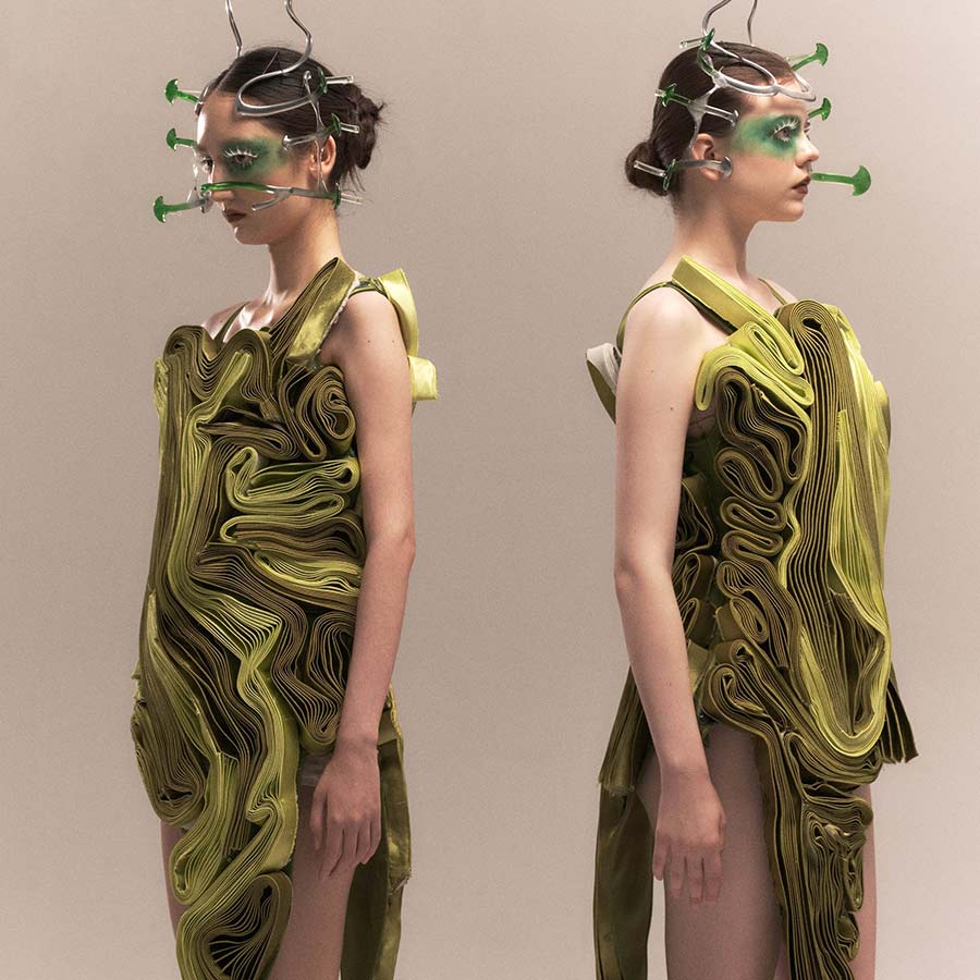 Double image of a model wearing a green, layered, sculpted dress with metal and green plastic facial ornamentation.