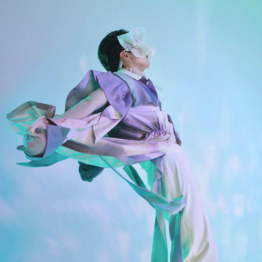A model wearing a light purple, white, and cream avant-garde outfit with a sculptural silhouette and a flower-like mask, against a blue and green gradient background.