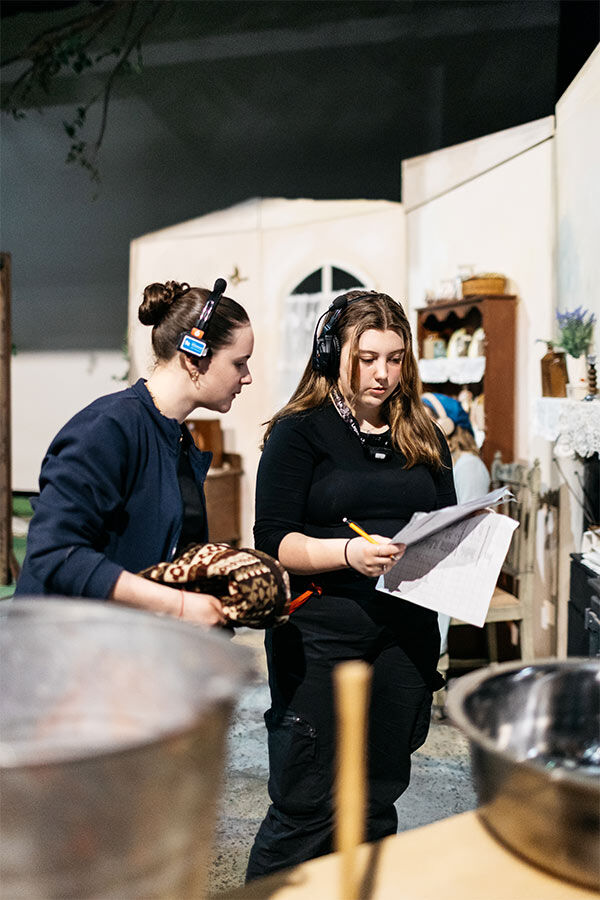 Two students are backstage, one is holding a prop and wearing headphones, the other is holding pieces of paper and wearing headphones