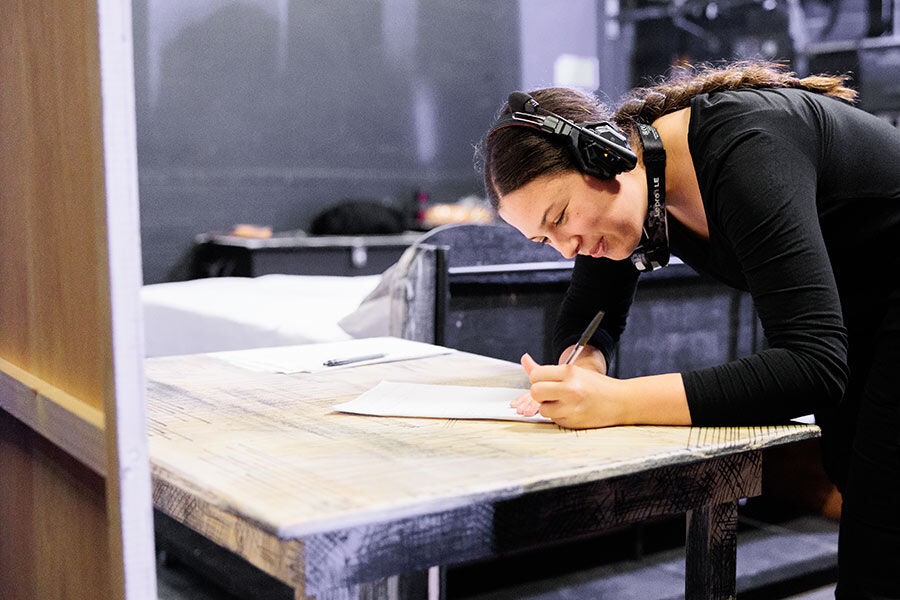 A student is writing on a piece of paper, wearing headphones, and is backstage 