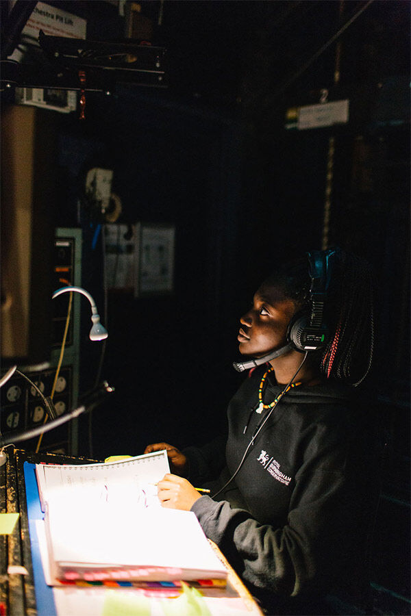 A student is working backstage in the lighting room, wearing headphones and looking at a screen