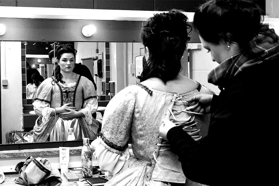 A black and white shot of a student getting ready to perform on stage, with another student working on her costume