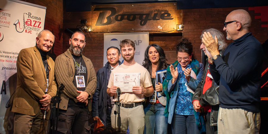 A group of people standing together on a small stage during an award ceremony at a jazz event. A young man at the center holds a certificate and smiles while others around him clap and celebrate. The background features a wooden wall with a sign that reads ‘Boogie’ and event banners.