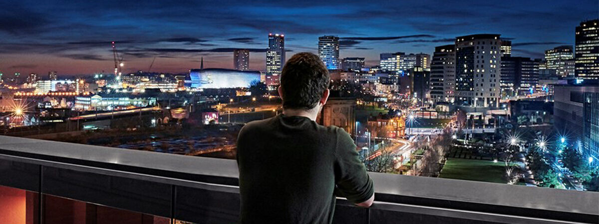 A person stands on a balcony overlooking a brightly lit Birmingham centre skyline at dusk or night.