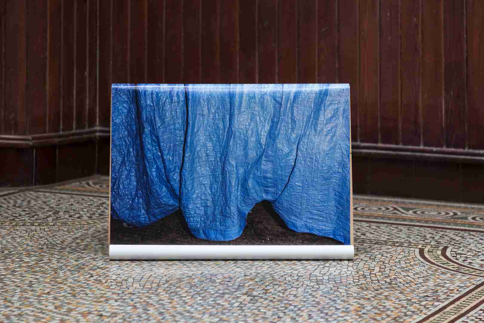 A blue draped fabric on a white stand, positioned against a dark panelled wall and a patterned floor