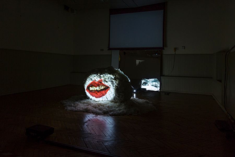 A brightly coloured projection of a face onto a textured, furry surface in a darkened room, with two video screens in the background
