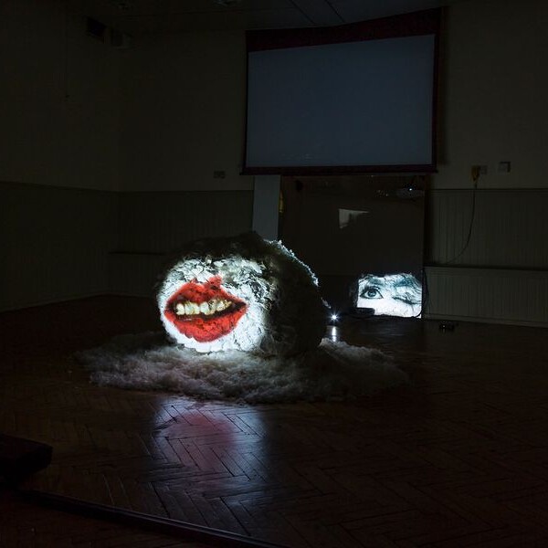 A brightly coloured projection of a face onto a textured, furry surface in a darkened room, with two video screens in the background