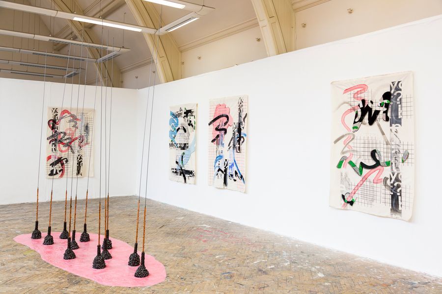 Gallery view of an art installation featuring three abstract paintings on the wall and a group of conical objects hanging from the ceiling over a pink shape on the floor
