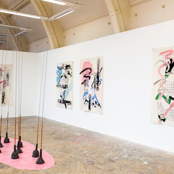 Gallery view of an art installation featuring three abstract paintings on the wall and a group of conical objects hanging from the ceiling over a pink shape on the floor