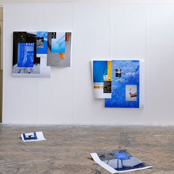 Gallery view of an art installation featuring three collage-like works on a white wall, with two additional pieces resting on the floor