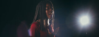 A person with long braids is singing with their eyes closed, illuminated by a bright light against a dark background.