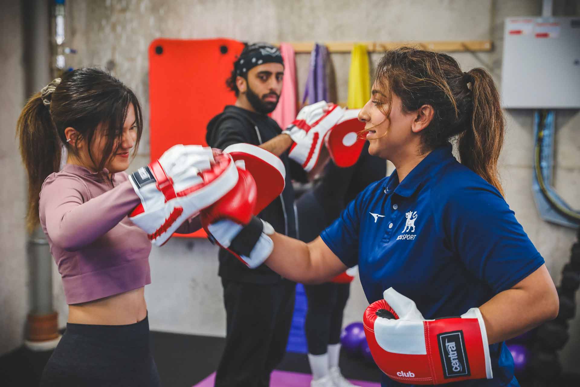 An instructor wearing boxing gloves and a BCU Sport shirt punches into boxing pads held by a student