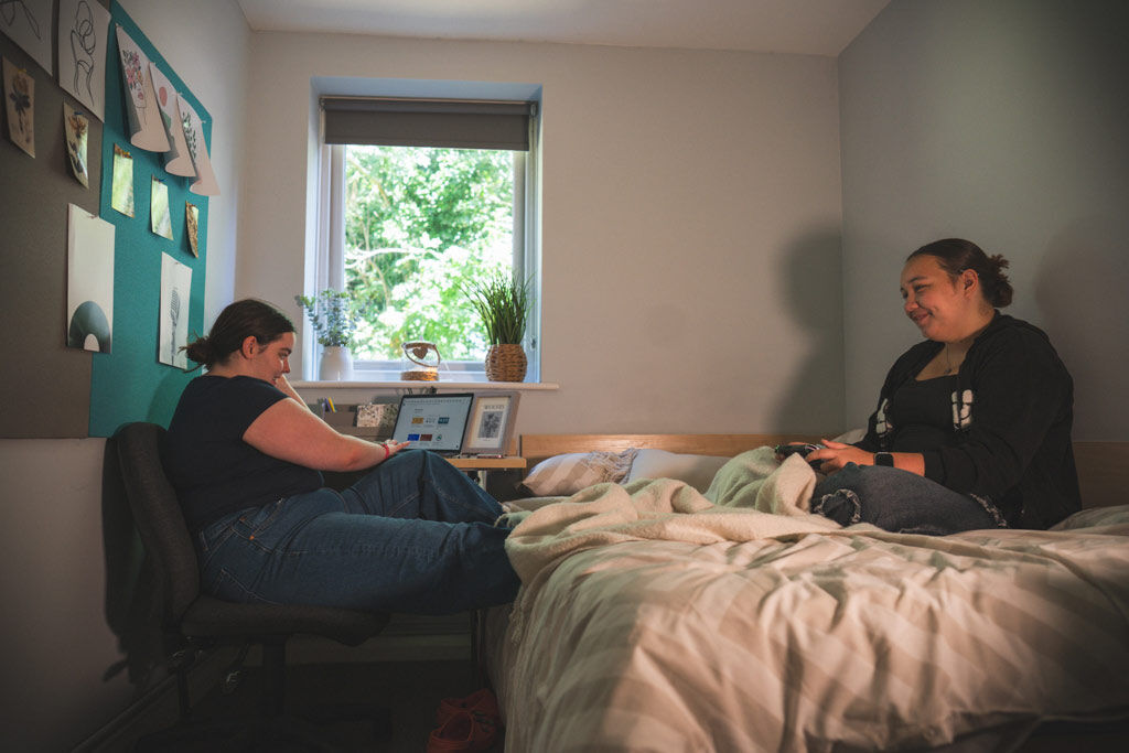Two students who appear to be female sitting in a bedroom. One is at the desk, one is sitting on the bed gaming, they look happy and relaxed