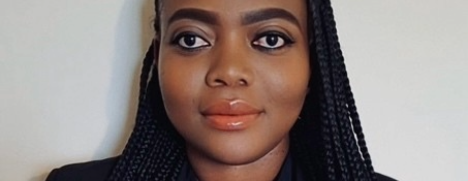 A close-up portrait of a person with neatly braided hair, facing the camera against a plain light background. They are wearing subtle eye makeup with defined eyebrows and eyeliner, and a soft peach-coloured lipstick. Their expression is calm and neutral, and they are wearing a dark top or blazer.
