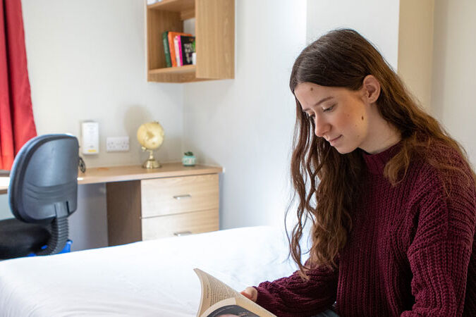 Student sits on her bed reading in her bedroom in university accommodation
