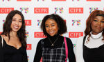 Three young women smile in front of a backdrop with "CIPR 75" logos. The woman on the left has long dark hair and wears a black top. The woman in the center has curly dark hair and wears a black turtleneck under a plaid dress. The woman on the right has long brown wavy hair and wears a black top over a white turtleneck.