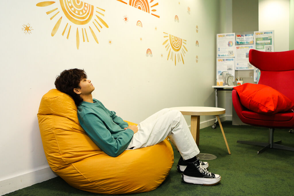A young person with light brown skin who appears to be male is sitting on a large yellow bean bag in a relaxed posture, leaning back with their hands resting on their stomach. They are wearing a teal long-sleeve sweatshirt, off-white trousers, and black high-top sneakers with thick white soles. Their eyes are closed, and they appear to be resting or meditating. The setting is a cozy indoor space with a green carpet, a white table, a red armchair with a matching cushion, and a wall decorated with orange sun-themed artwork.