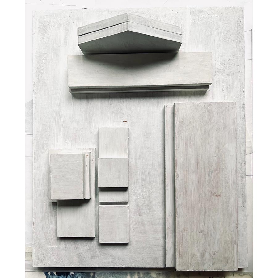 A grayscale photograph of a minimalist abstract sculpture. Stacked wooden blocks of varying sizes and shapes, arranged in a geometric composition against a textured background