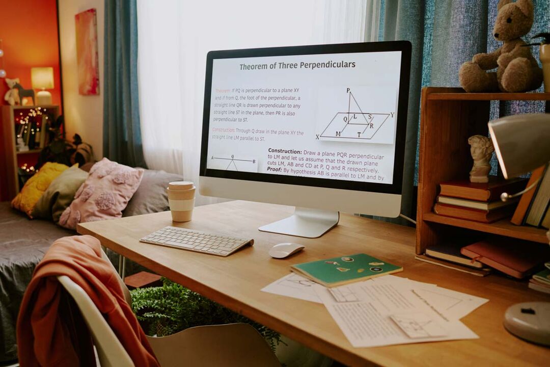 A modern desk setup in a cosy bedroom features a large computer monitor displaying a geometry lesson on "Theorem of Three Perpendiculars." The desk holds a wireless keyboard, mouse, a takeaway coffee cup, notebooks, and loose papers with diagrams. To the right, a wooden shelf contains books, a small lamp, and a plush teddy bear. In the background, a bed with colourful cushions and a blanket sits against an orange accent wall, with a string of fairy lights adding a warm touch. A window with light blue curtains lets in natural daylight, creating a calm and focused study atmosphere.