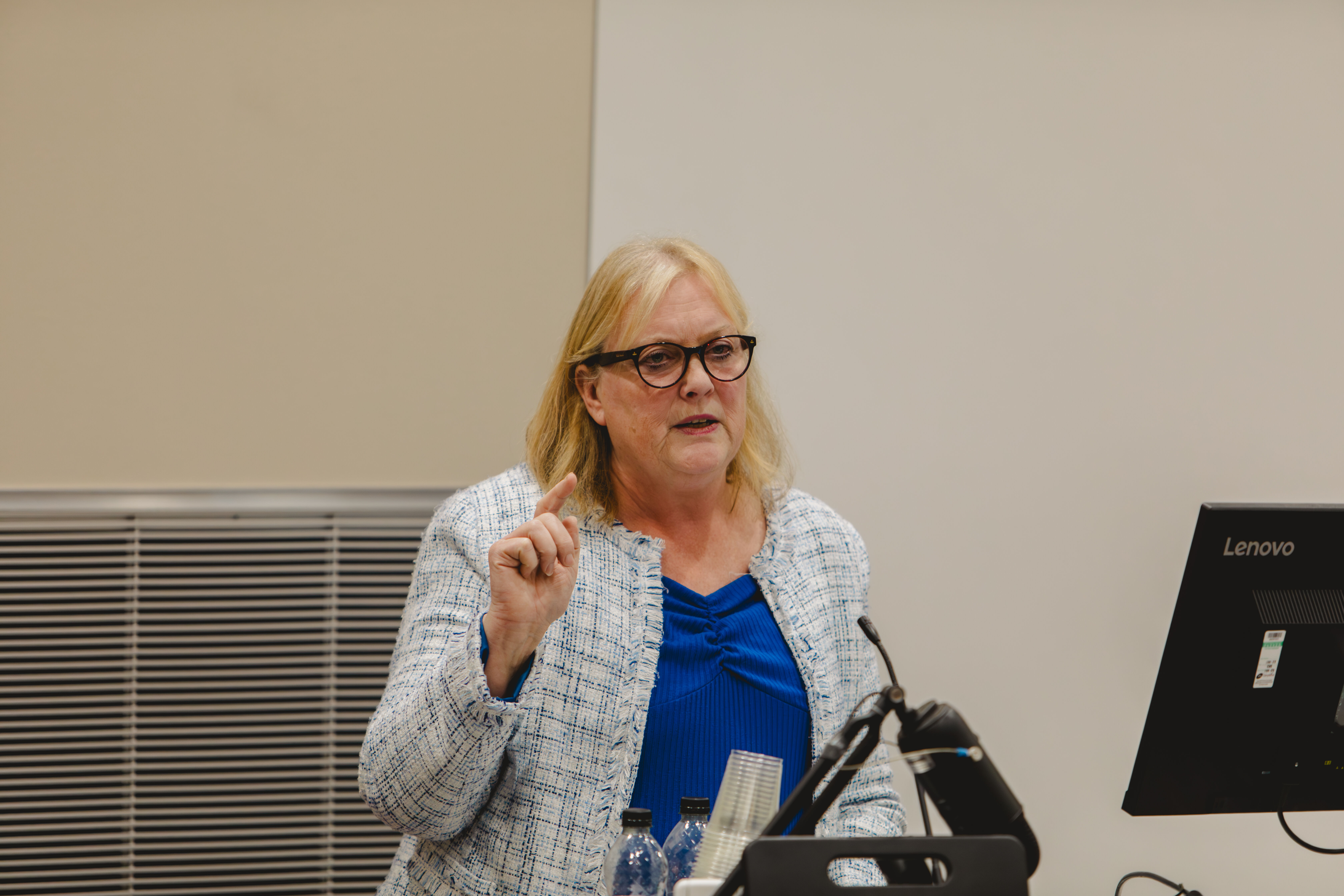 A lady with blonde hair and glasses points to an audience and presents