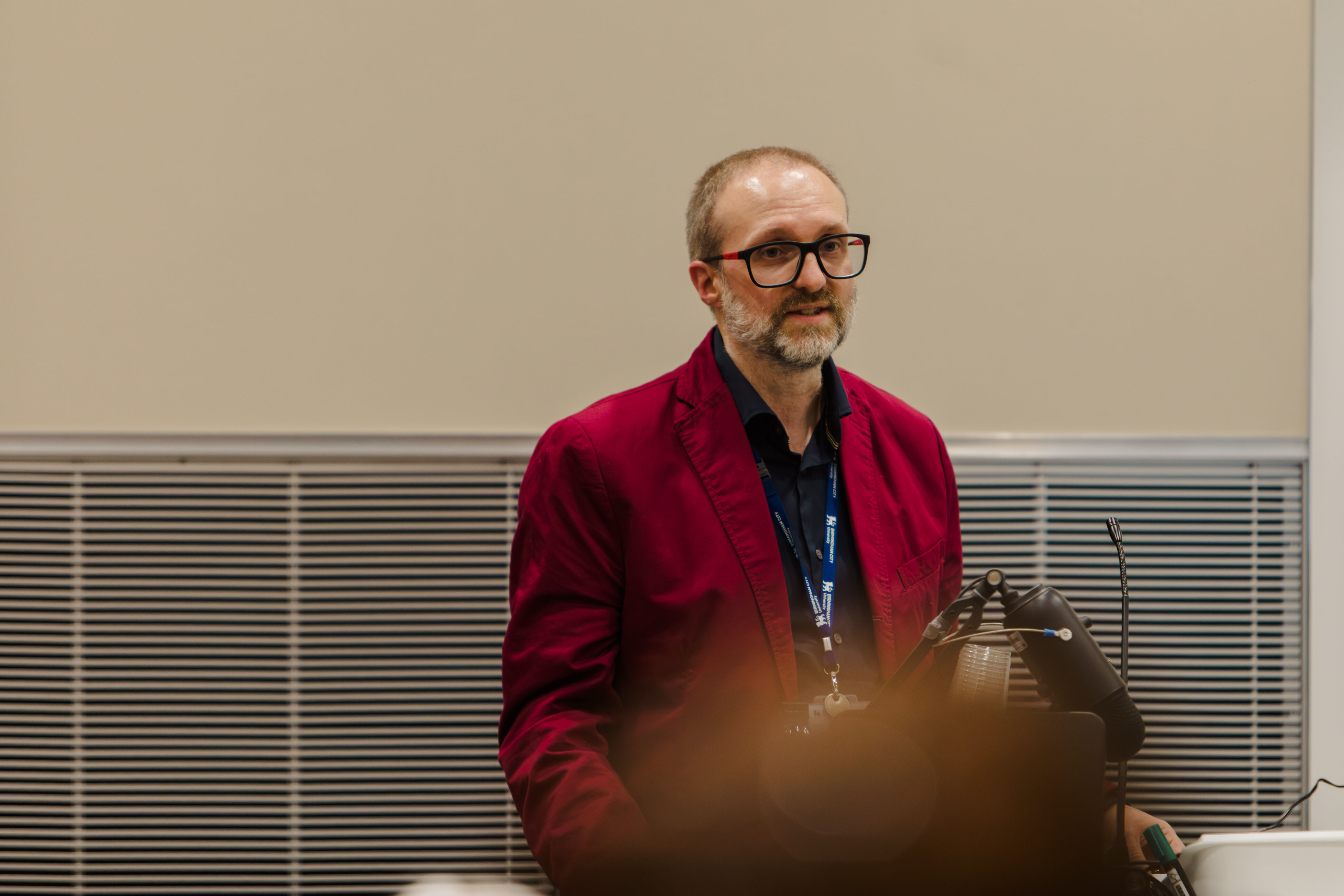 A man in a dark red blazer and glasses looks at the audience