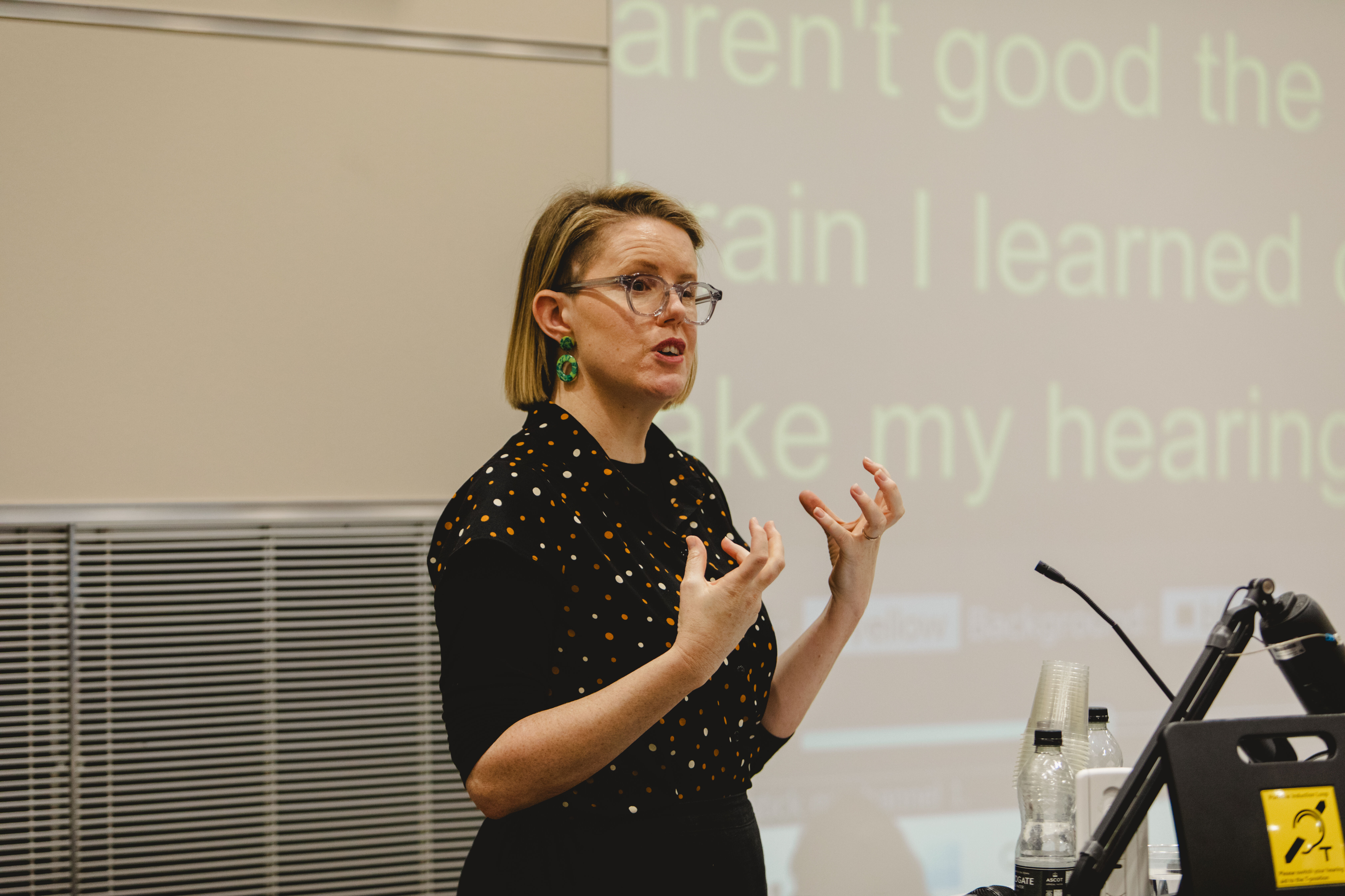 A lady with blonde hair and glasses talks to the audience with her hands