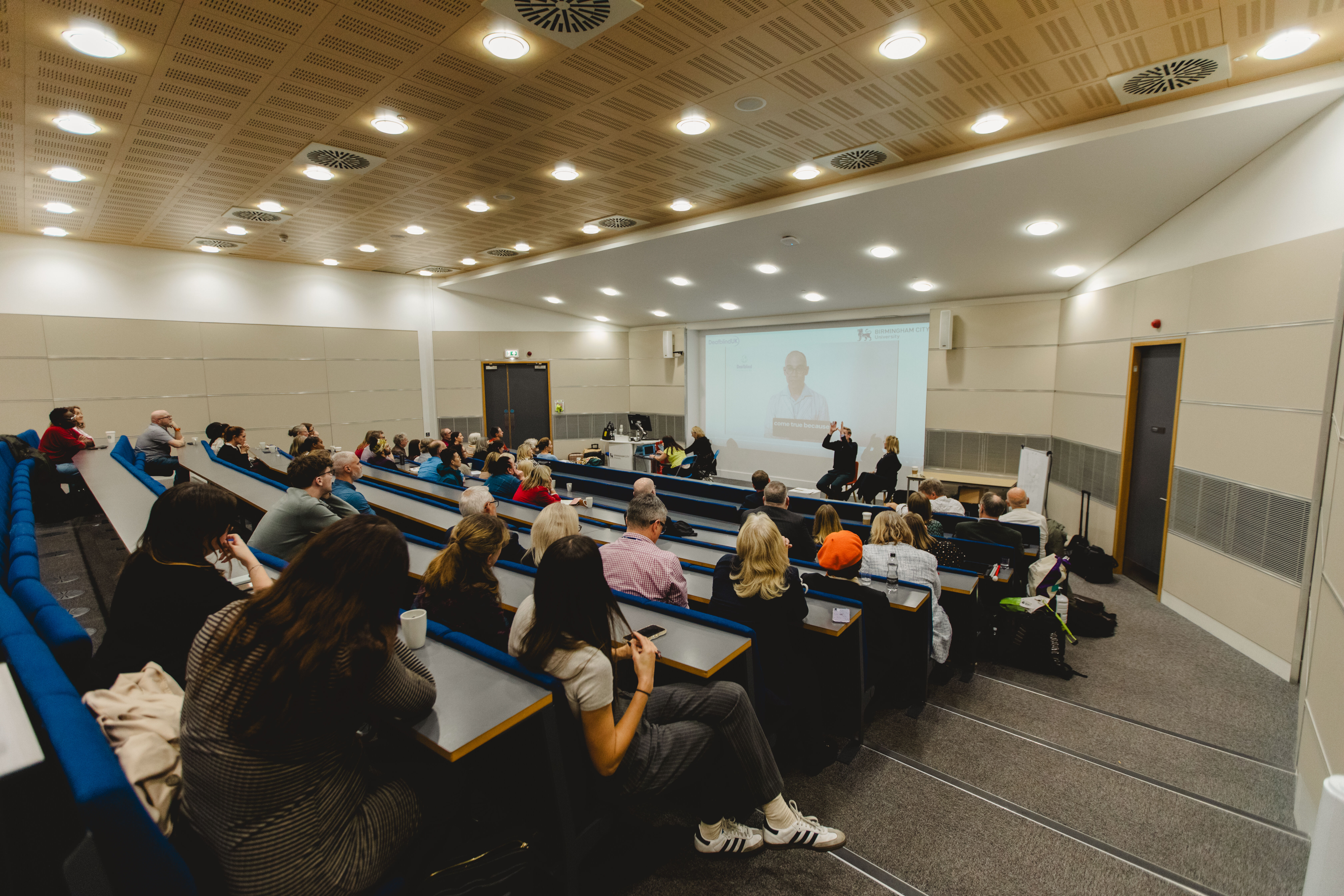 A warm lecture theatre with people sat facing a man signing