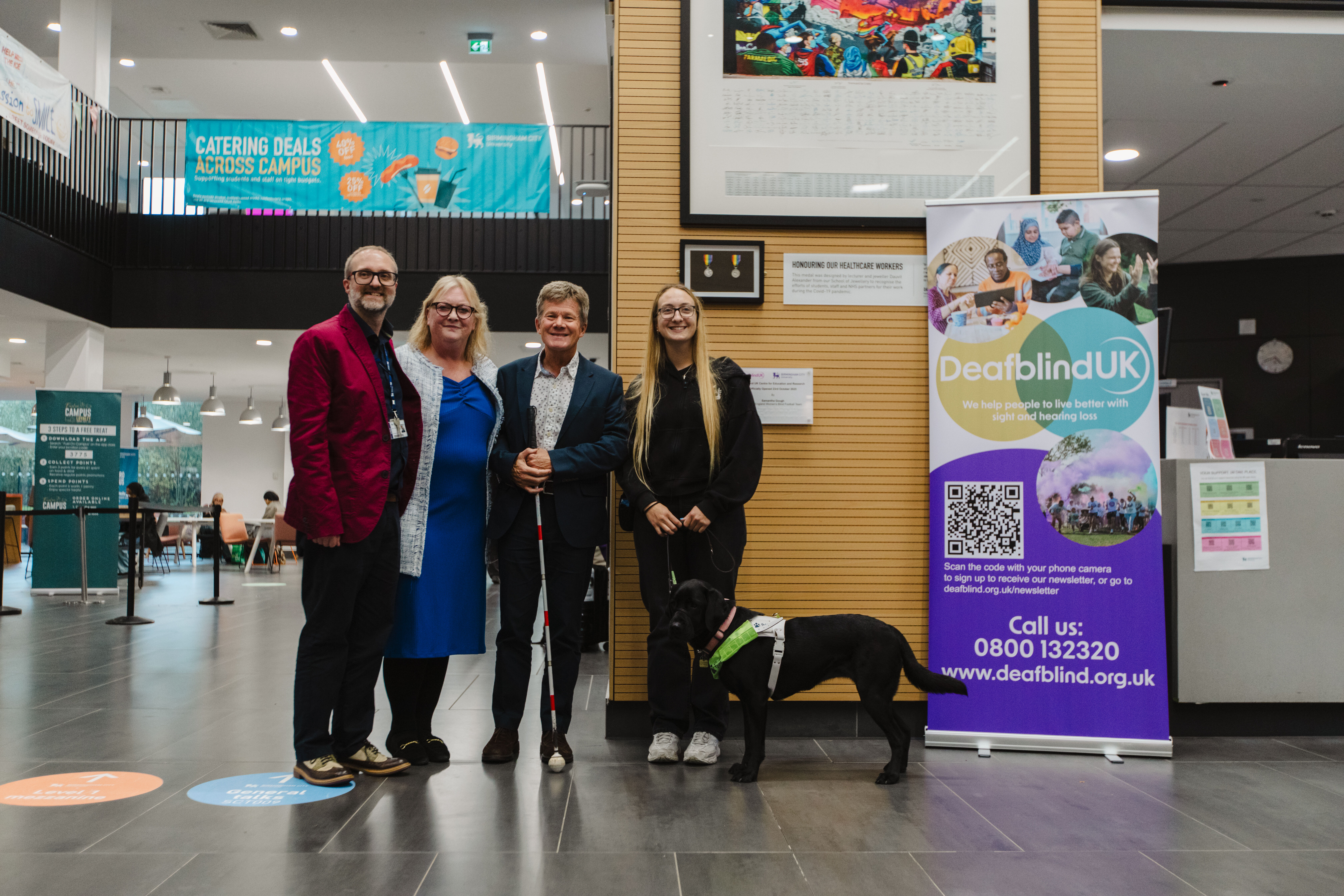 Four people, two male and two female and a guide dog stand in front of a sign reading "Deafblind UK" smiling