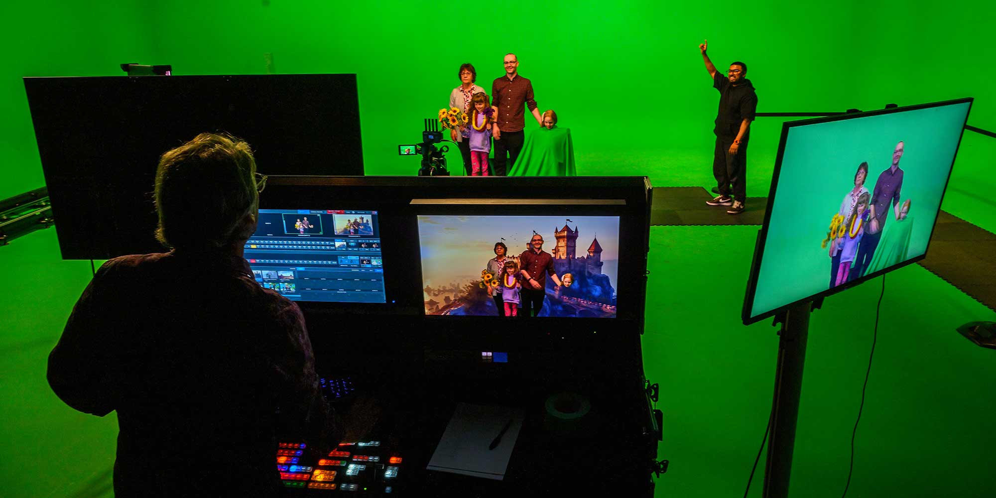 A family having fun taking photos on the green screen. They are standing against a green backdrop with props, in the foreground a person operates the screen and the family are shown in front of a castle.