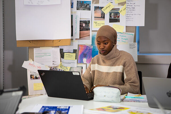 A young woman is working hard on her laptop at a desk. There are lots of papers and notes pinned up behind her – looks like she's got a big project on the go!
