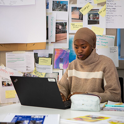A young woman is working hard on her laptop at a desk. There are lots of papers and notes pinned up behind her – looks like she's got a big project on the go!