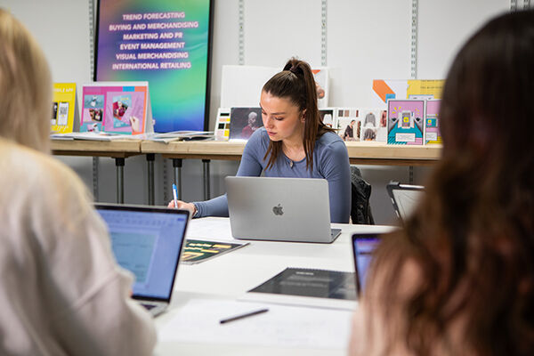 A young woman with her hair in a ponytail is working on a silver laptop and writing on some papers. She looks focused on her work! There are other people and laptops around the table too. It seems like a study group or a class!