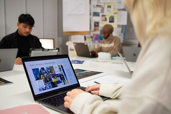 Three young people are working on their laptops around a table. The person in the foreground is looking at a website with pictures of clothes. It looks like they might be doing some research or working on a project together!