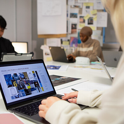 Three young people are working on their laptops around a table. The person in the foreground is looking at a website with pictures of clothes. It looks like they might be doing some research or working on a project together!