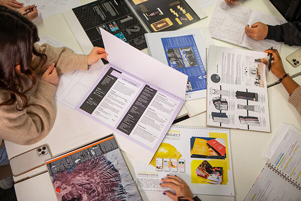 A group of young people are sitting around a table, looking at different papers and notebooks. It looks like they're working on a school project together!
