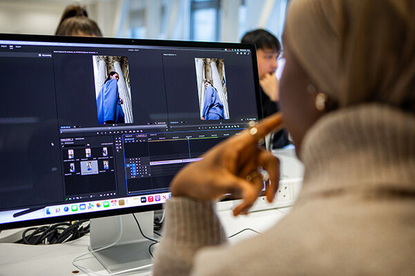 Someone is working on a big computer screen, editing a video of a person wearing a cool blue outfit! It looks like they're putting together something really interesting. There are other people in the background watching too!