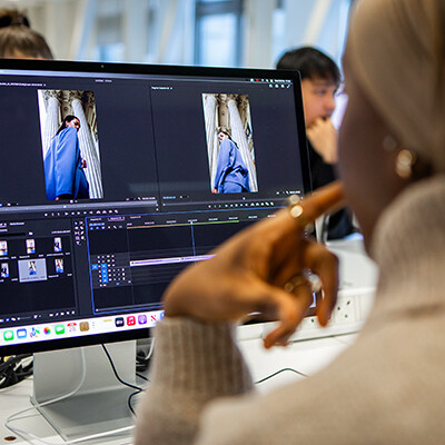 Someone is working on a big computer screen, editing a video of a person wearing a cool blue outfit! It looks like they're putting together something really interesting. There are other people in the background watching too!