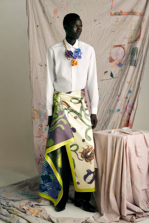 Male model in a white shirt, a vibrant, draped botanical-print satin sarong skirt, and a colourful statement necklace, standing against a paint-splattered fabric backdrop.