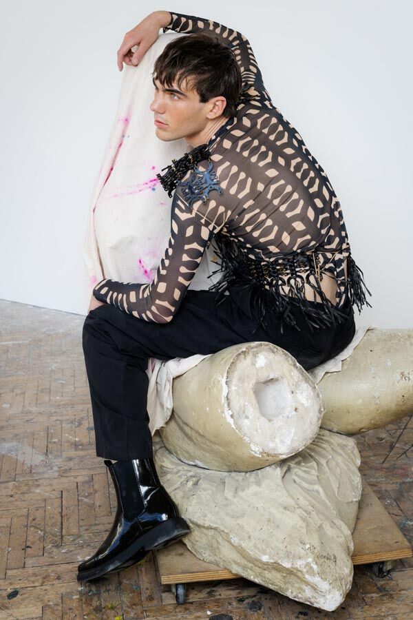 Male model in a patterned, sheer long-sleeve top with fringed details, black trousers, and shiny black boots, sitting on a large stone architectural fragment.