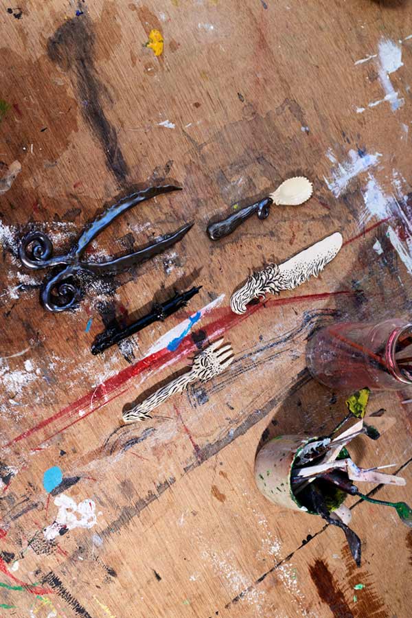 Top-down view of ornate, artistically sculpted utensils—including a fork, knife, spoon, and tongs—laid out on a paint-splattered wooden workbench next to cups of paintbrushes.