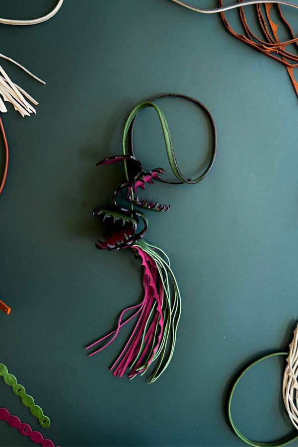 Top-down view of a sculpted leather necklace shaped like open Venus flytraps with long pink and green fringe, displayed on a dark green surface surrounded by material scraps.