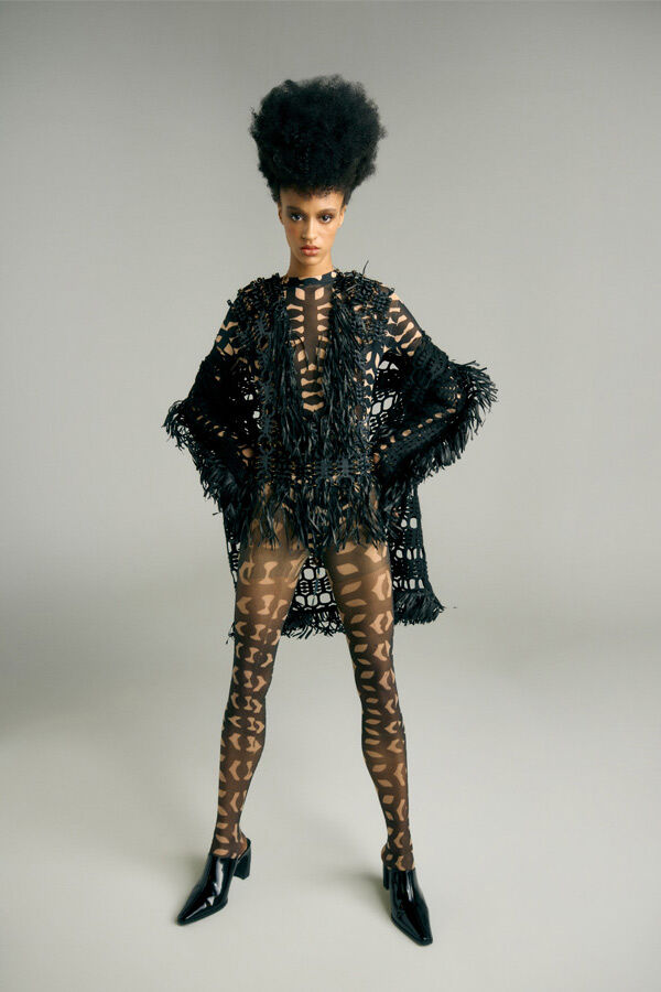 Full-length fashion portrait of a female model with a high afro hairstyle, wearing a patterned, cut-out bodysuit and matching tights under a heavy, fringed black crochet jacket, standing against a plain grey background.